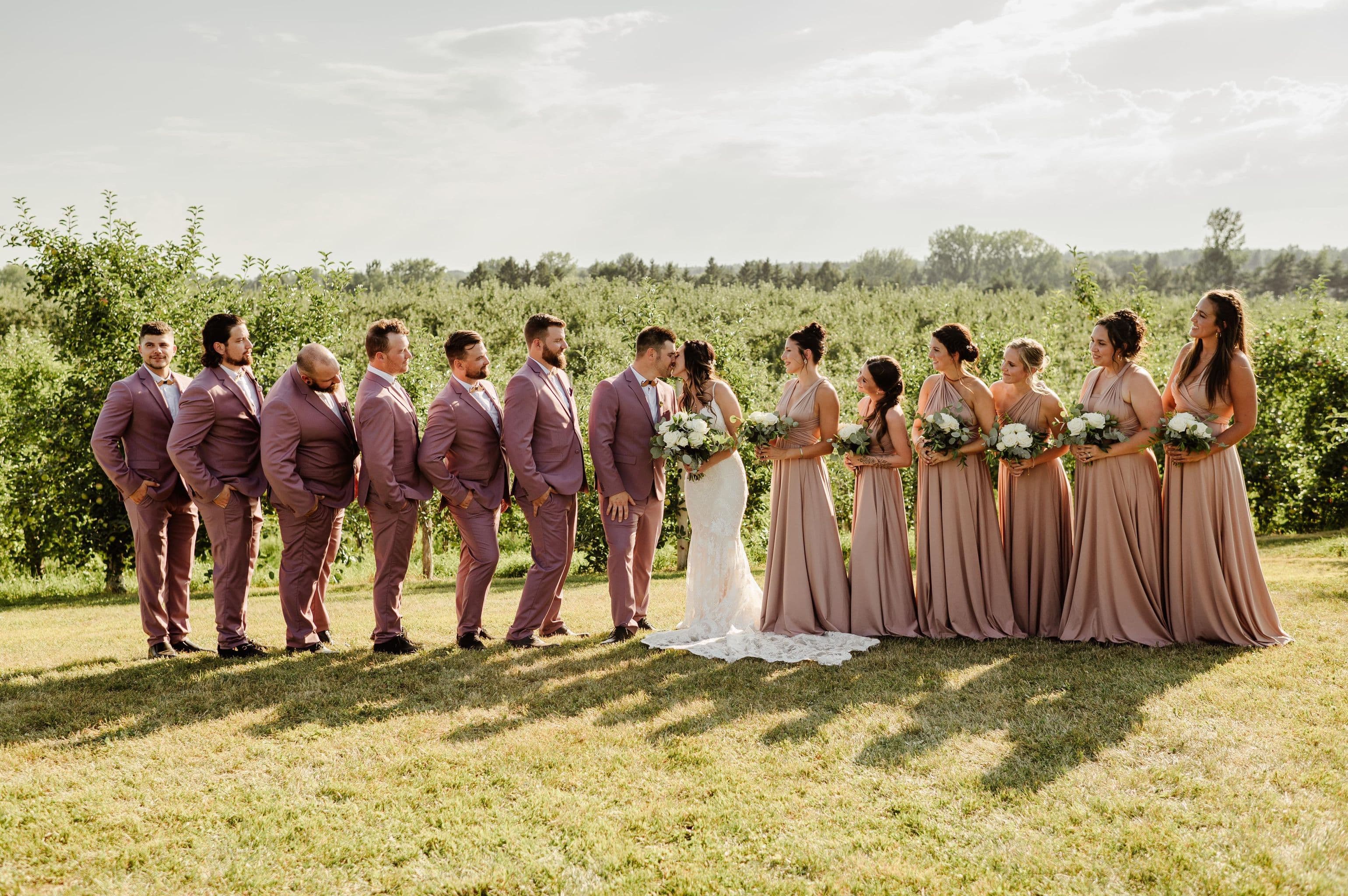 Cortège de mariage aligné dans le verger, costumes mauves et robes dusty rose