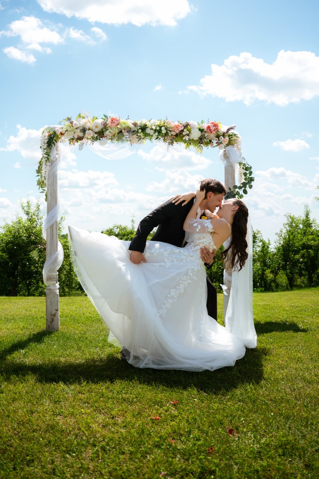 Newlyweds kissing under the floral arch, blue sky and orchard in the background